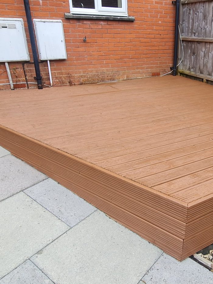Newly laid brown decking outside a brick house, adjacent to a paved area.
