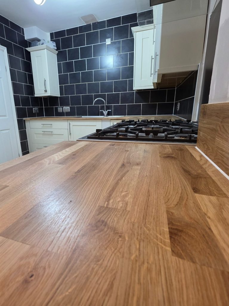 Oak kitchen worktop in a modern L-shaped kitchen with black wall tiles and white wall hung cabinets.