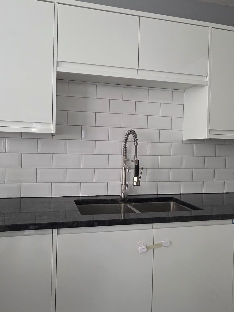 Subway style white ceramic tiles laid along a countertop behind a kitchen sink as a backsplash with light grey grout between.