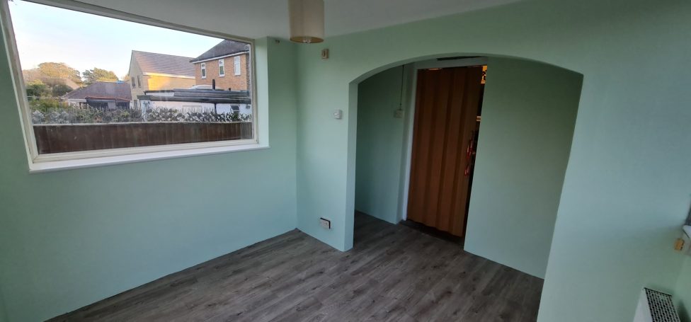 Empty room with green walls, wooden flooring, and an arched doorway. Natural light from window.