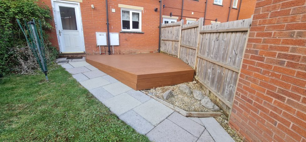 Brown decking area next to a paved path in a garden with wooden fencing.