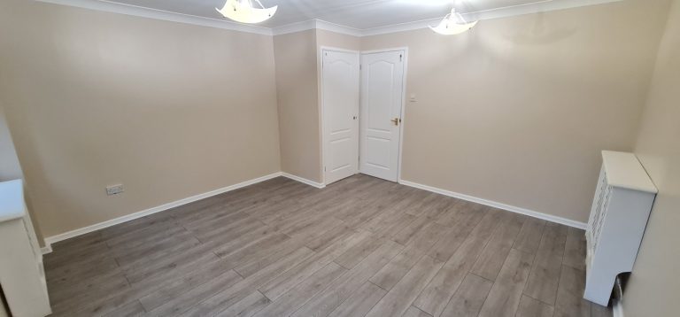 Empty room with beige walls, wooden floor, and a white door. Two ceiling lights visible.